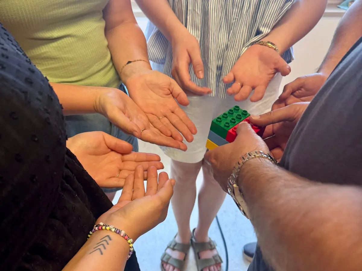 Sechs bunte Steine in der Hand, umgeben von offenen Händen im Kreis